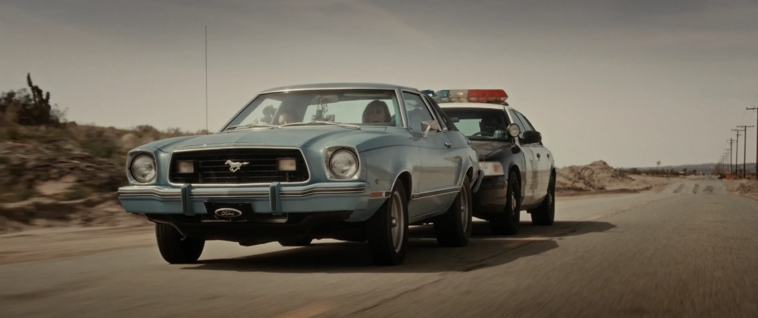 A sheriff's patrol car pursues Bobbi's Mustang on an empty desert highway in Blood Star