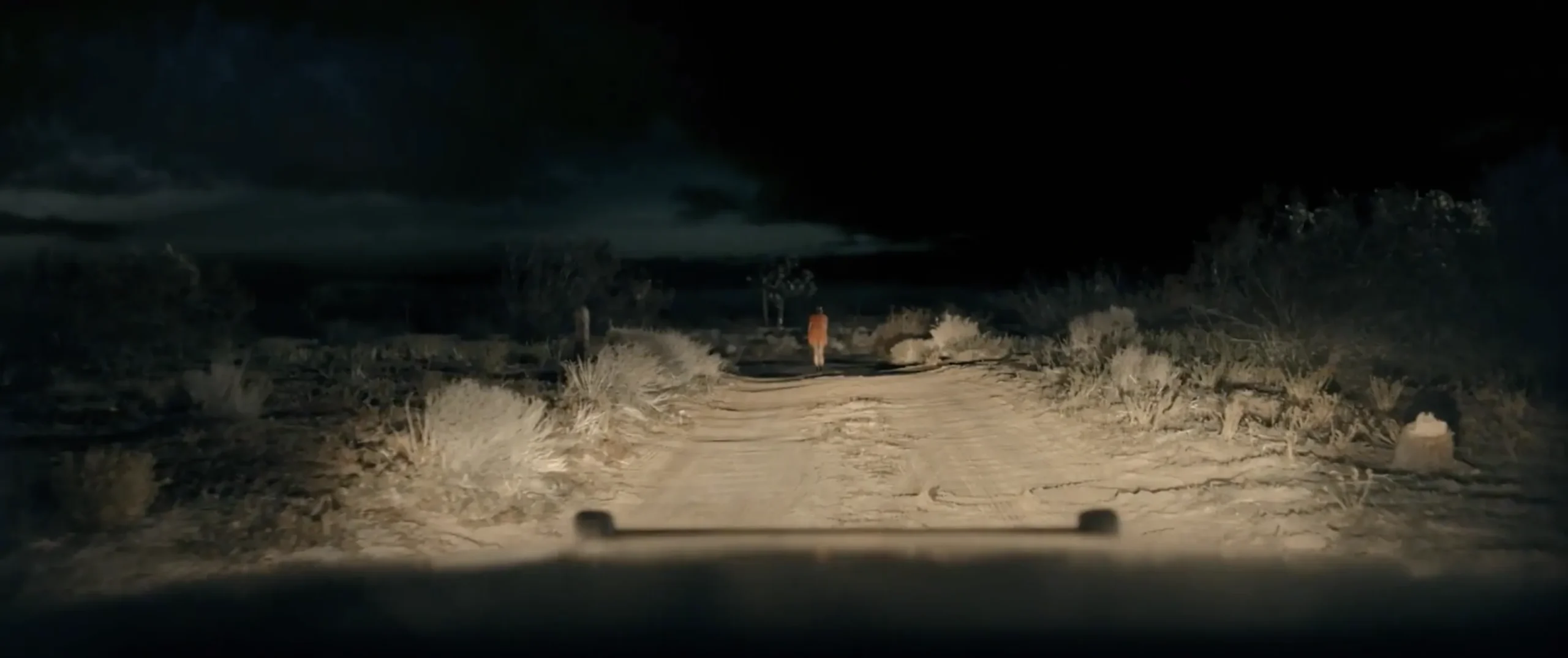 A dark desert road seen from behind a vehicle's headlights, with a lone figure walking in the distance