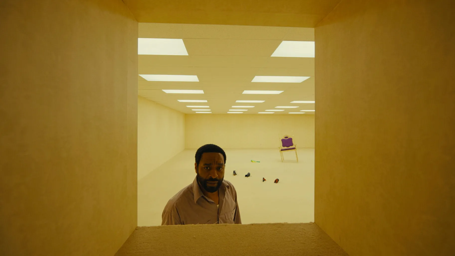 A vast empty room seen through a doorway, with a single purple chair and scattered belongings on the floor