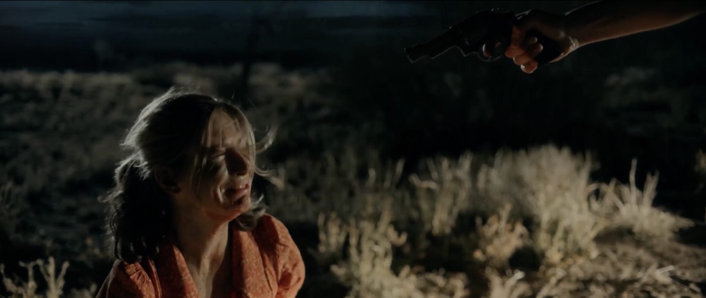 A revolver held out toward a bloodied young woman kneeling in the dark desert, Blood Star's opening ritual