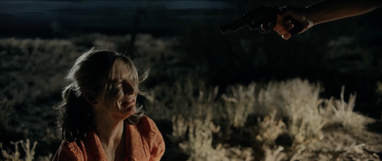 A revolver held out toward a bloodied young woman kneeling in the dark desert, Blood Star's opening ritual