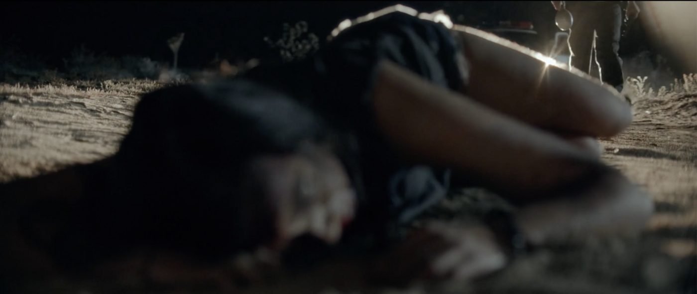 A bloodied woman lying in the dark desert as the sheriff approaches in the background