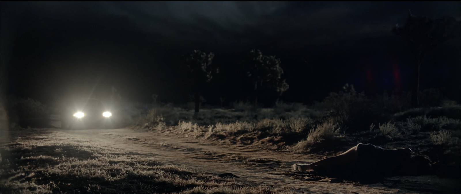 Wide shot of Bilstein's cruiser headlights approaching down a desert road with a body lying in the foreground