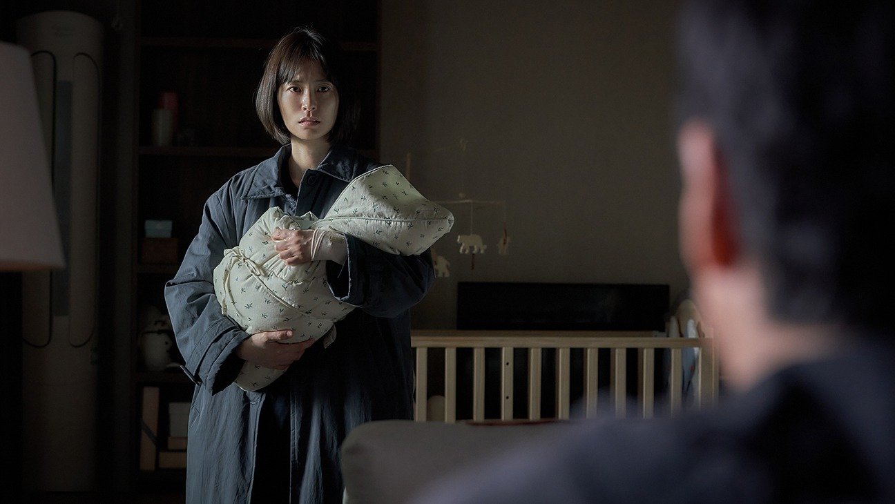 A woman holding a swaddled baby while a man's silhouette watches from the foreground in Sleep (2023)