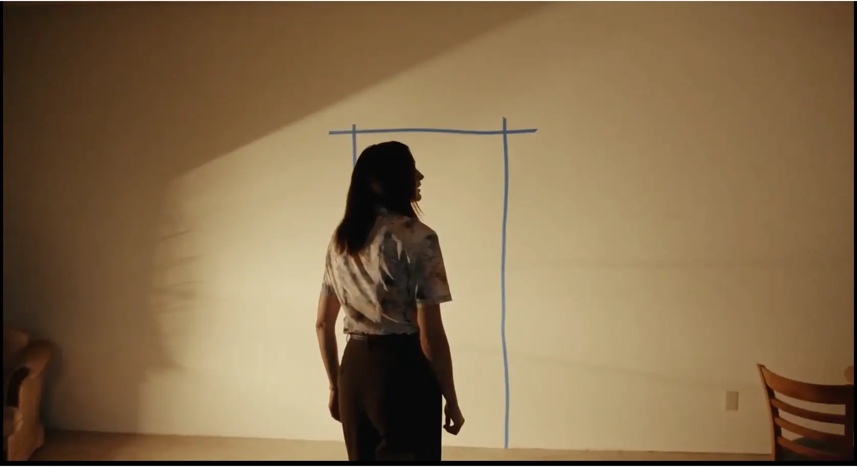 A character seen from behind, standing before a doorway outline drawn in tape on a blank wall