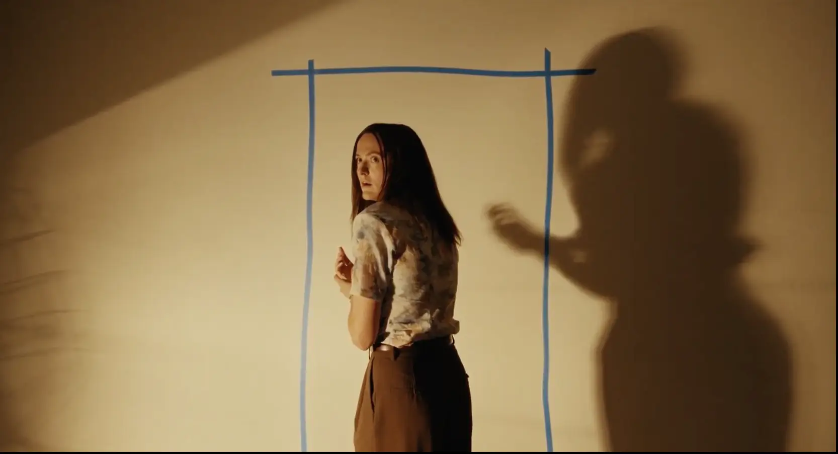 A character stands inside a doorway outline drawn in tape on a blank wall, with a huge shadow looming beside her