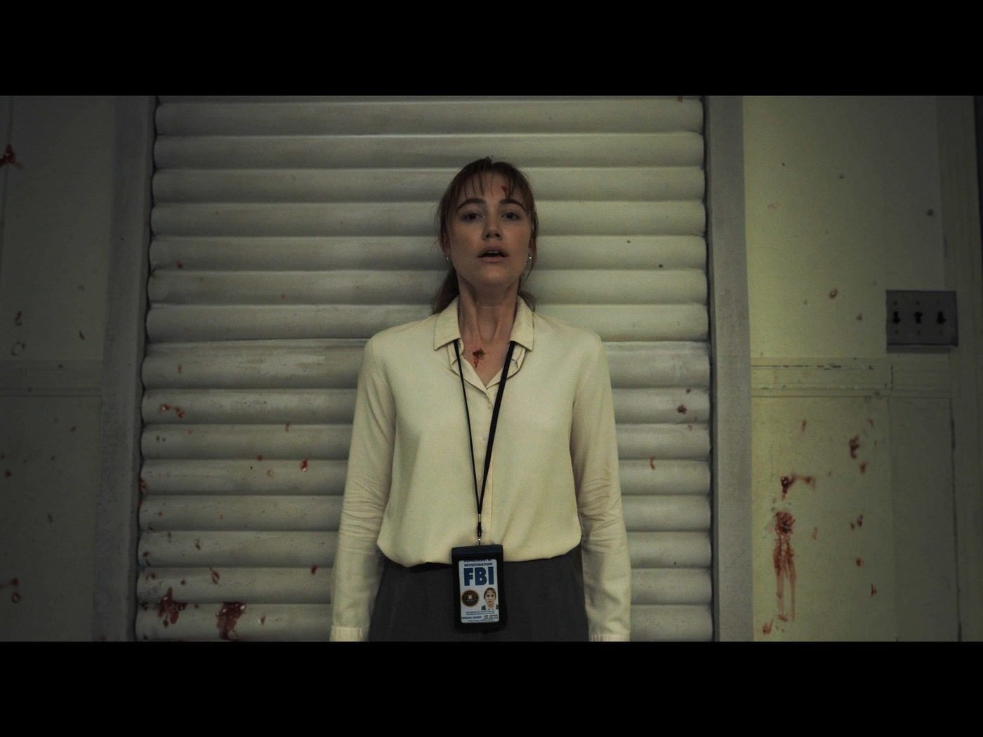 Agent Lee Harker, bloodied, wearing an FBI badge, standing against a wall spattered with blood in Longlegs (2024)
