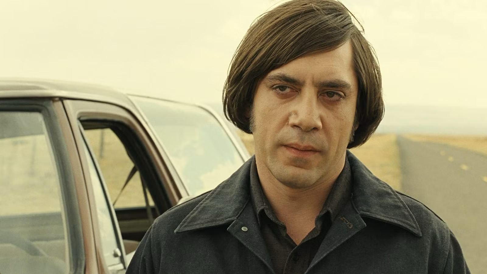 Anton Chigurh standing beside a truck on a rural road in No Country for Old Men